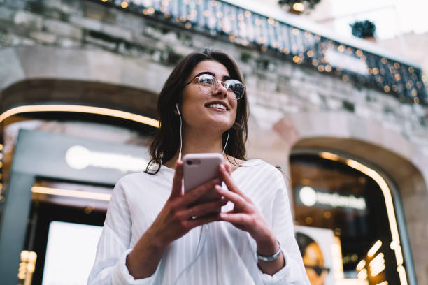 gioiosa utente femminile in auricolari digitali che si godono canzoni musicali durante il tempo libero del passatempo in città, allegra ragazza hipster in occhiali ottici per la protezione della vista ascoltando dischi audio e sorridendo - spanish culture audio foto e immagini stock