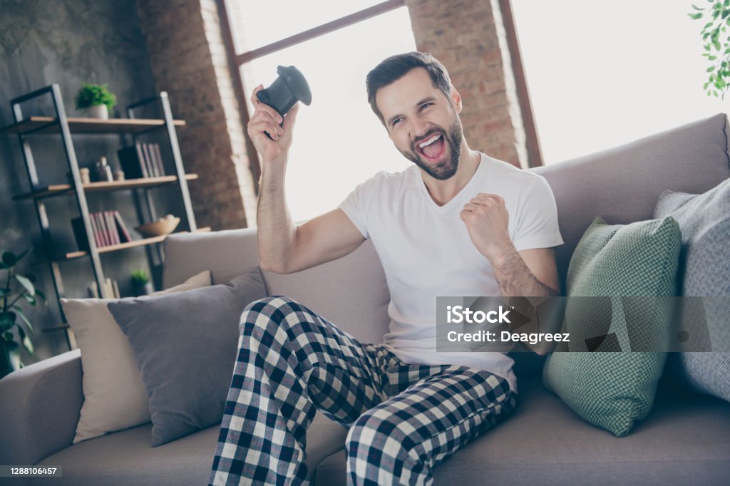Photo of handsome excited guy homey sit comfy sofa quarantine stay home gamer hold play station joystick good mood fifa championship pajama pants t-shirt living room indoors Photo of handsome excited guy homey sit comfy sofa quarantine stay, home gamer hold play station joystick good mood fifa championship pajama pants t-shirt living room indoors Domestic Life Stock Photo