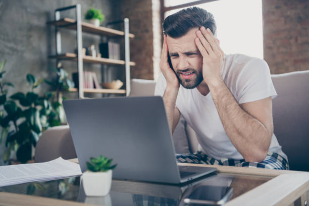 istock Photo of handsome focused serious tired guy sit sofa work morning overwhelmed loaded browsing notebook freelancer remote work home quarantine read contract pajama living room indoors 1288107792