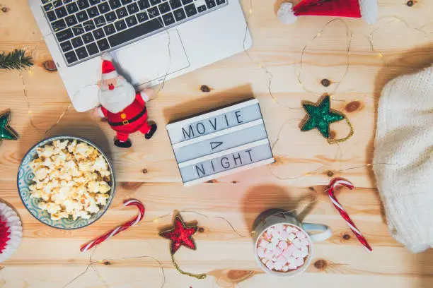 Top view Christmas Movie night concept. Flat lay composition with Movie night message on the board, laptop, popcorn bowl, decor, a cup of cocoa with marshmallows and warm plaid on wooden background
