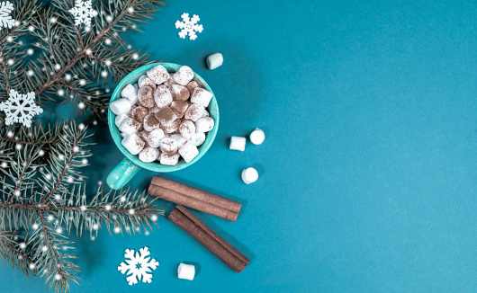 Bright turquoise Christmas background with fir branches with snow and cup of coffee or cocoa with marshmallows. Cozy home holiday. Xmas decorations. Top view, flat lay. Copy space for text.