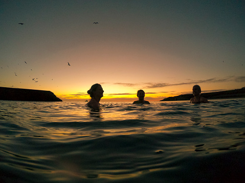 A wide angle shot of a group of three female friends swimming in the North Sea during the early morning in Autumn. They are talking and bonding, enjoying their time swimming together.