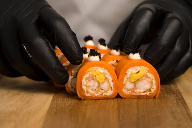 fermez-vous vers le haut aux mains de chef dans les gants protecteurs noirs près du japonais aucun rouleau de riz enveloppé dans le daikon de carotte. le chef tient des sushis. natures sans vie - carrot close up silverware cutting board photos et images de collection