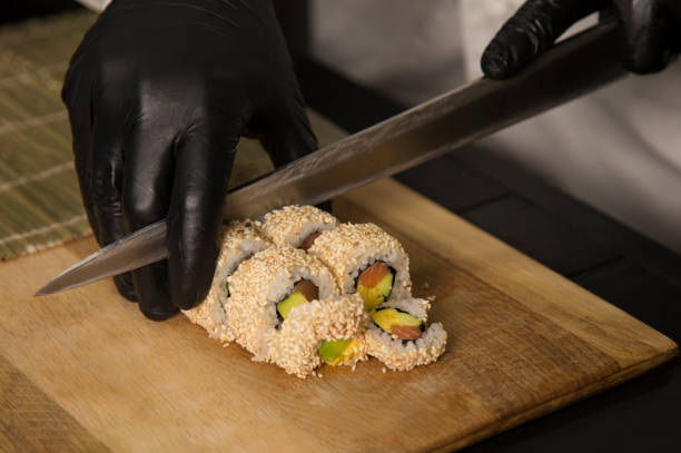 szef kuchni tnie japońską rolkę nożem w rękawicach ochronnych. profesjonalny proces gotowania cięcia sushi roll z łososiem, awokado zawinięte w ryż z nasion sezamu. - 3693 zdjęcia i obrazy z banku zdjęć