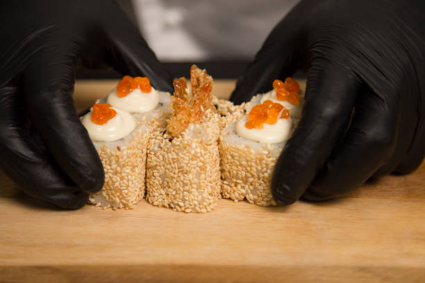 chef’s hands in protective gloves near japanese ebi tempura sushi roll with black tiger shrimp inside. man holds food in hands. serving process in restaurant kitchen - 3679 imagens e fotografias de stock