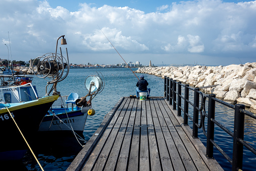 April 11, 2019, Larnaca, Cyprus. A fisherman sits with a fishing rod on a pier behind a stone breakwater on the embankment.
