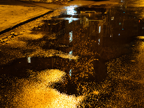 reflection of glowing windows of residential city house in rain puddle on autumn night