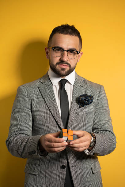istock Smart young bearded handsome man in eye glasses wearing a suite holds a solved Pocket Cube 2x2x2 rotatable puzzle in hands while showing it on camera. Isolated on yellow background 1288127093