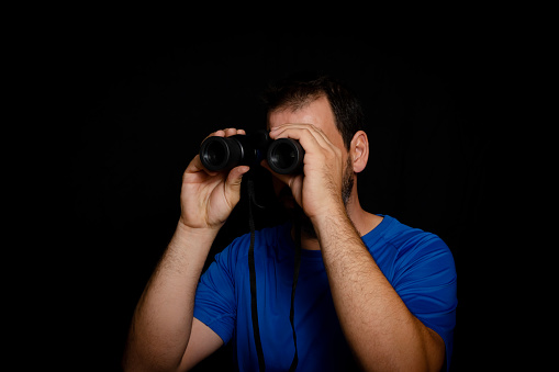 Bearded man dressed in blue t-shirt with binoculars posing against black background. Fashion concept