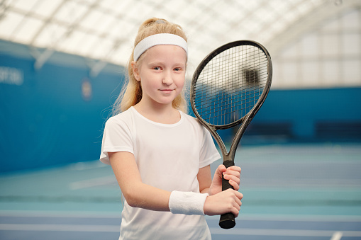 Cute blond little girl in white activewear holding tennis racket by left shoulder while standing in front of camera in stadium environment