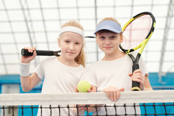 肩にテニスラケットを保持している白いスポーツウェアで2かわいいフレンドリーな女の子 - tennis indoors women court ストックフォトと画像