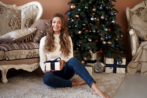 Christmas. Woman dressed white sweater and jeans sitting on the floor near christmas tree with present box Caucasian female middle age opens gifts winter holiday at home interior New year mood
