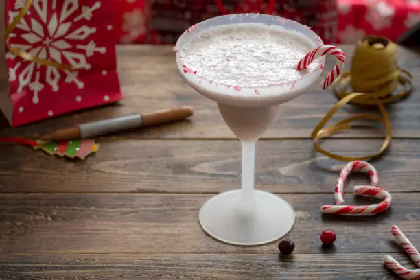 Candy cane Christmas Smoothie. Frosted style cocktail glass filled with creamy smoothie and a candy cane with candy cane crumbles on the rim. Gift wrap, label and ribbon soft focus in the background