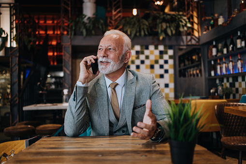 Happy and handsome senior businessman sitting in restaurant and waiting for lunch. He is using smart phone and talking with someone. Business seniors lifestyle concept.