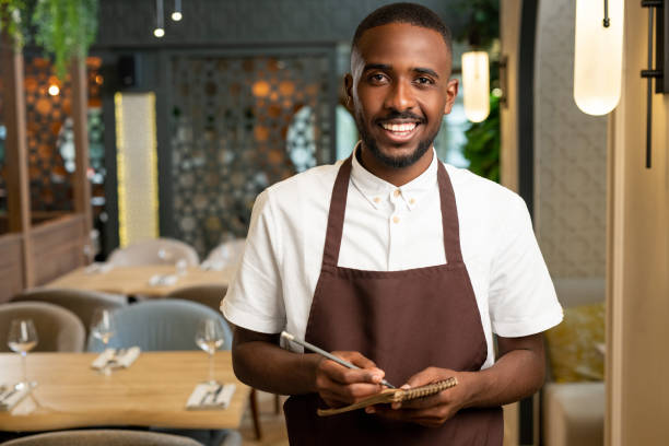 istock Happy young waiter with toothy smile taking orders with pen and small notepad 1288149634
