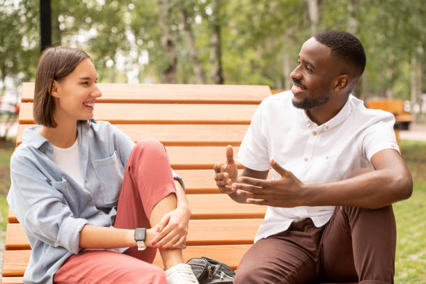 Happy young intercultural couple in casualwear sitting on wooden bench in park Happy young intercultural couple in casualwear sitting on wooden bench in park or square, chatting and enjoying summer day at leisure black man white woman couple stock pictures, royalty-free photos & images