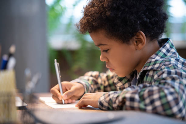 istock Side view of cute little African schoolboy making notes in copybook by table 1288153914