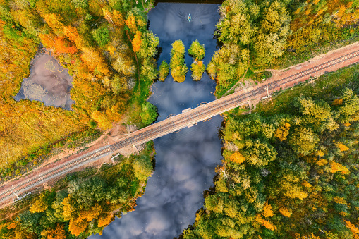 Forest river with sky reflection crossed by railroad. Aerial top view in autumn colors