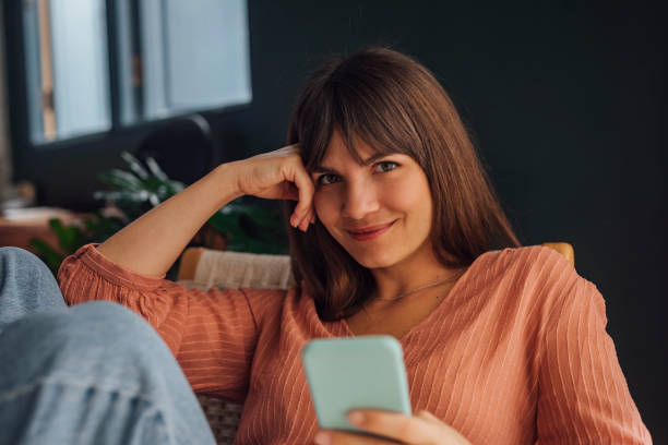 Beautiful Young Woman Looking at Camera and Using a Mobile Phone Sitting in a Chair Beautiful cheerful young Caucasian woman wearing a pink blouse and jeans looking at camera using a mobile phone while sitting casually in a chair. 30 34 years stock pictures, royalty-free photos & images