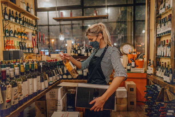 Woman packing wine bottles in shipping boxes Photo of a millennial woman wearing a protective face mask while packing wine bottles into a delivery box; running a wine shop and working during a disease outbreak. wine shipping stock pictures, royalty-free photos & images