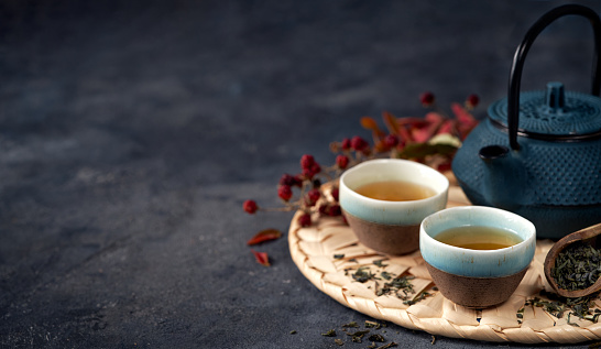 Asian food background banner with green tea, cups and teapot with leaves and free space for text on dark background.