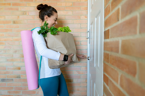 Woman arriving home with the groceries after her workout and carrying her yoga mat while opening the door - lifestyle concepts