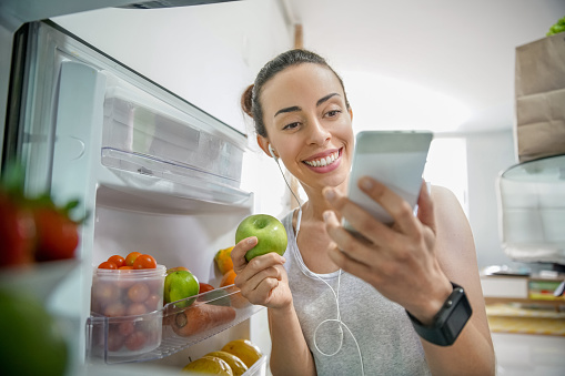Portrait of a happy Latin American woman at home grabbing food from the fridge while watching videos online on her cell phone and using earphones
