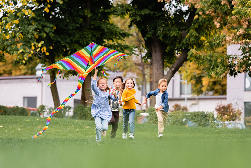 Smiling multiethnic kids running on lawn while playing with flying kite in park