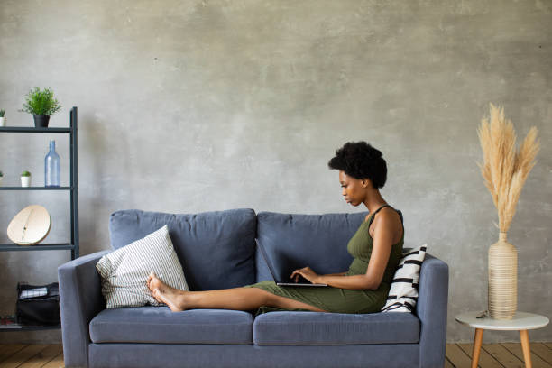 Happy african American young woman sit relax on cozy couch and working on a laptop. Happy to move to new apartment stock photo