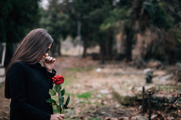 Young woman grieves in a cemetery holding rose One young sad woman, widow, visiting a loved one at the cemetery. Paying respect with fresh rose flower. widow stock pictures, royalty-free photos & images