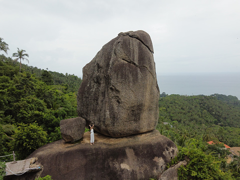 Aerial view of woman, Female traveller wear white dress on the edge of the huge rock over beautiful green mountains, palm trees and sky background at Koh Samui Island, Southeast Asia, Thailand