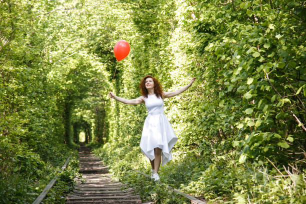 Young woman with red balloon stands on rail of railway on forest background