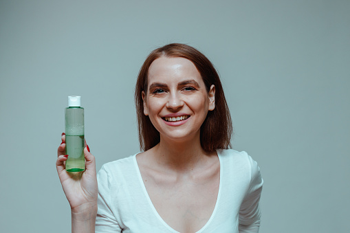 Portrait of a smiling young woman holding bottle with a beauty product micellar water against a bright blue background