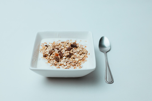 Healthy cereal muesli with oats and raisins in a squared shaped white bowl and a silver spoon against a bright blue background