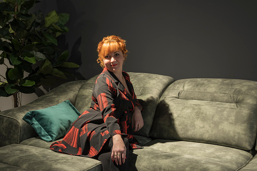 Young red-haired girl sitting on the sofa half-turned on the sofa in a dark room