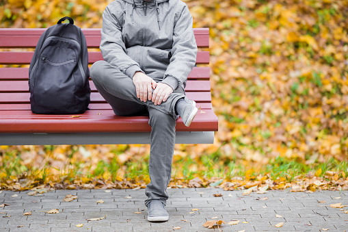 One young adult man in gray clothes sitting on wooden bench at park in autumn day. Front view. Close up. Black backpack beside guy.