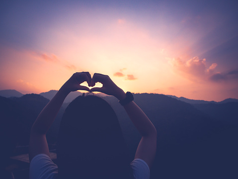 silhouette of young woman making heart shape with hands in the atmosphere sunset on mountain peak.