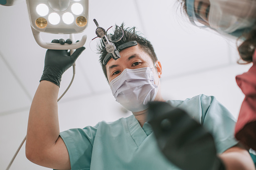 asian chinese male dentist with magnifying glasses and light looking from above at patient