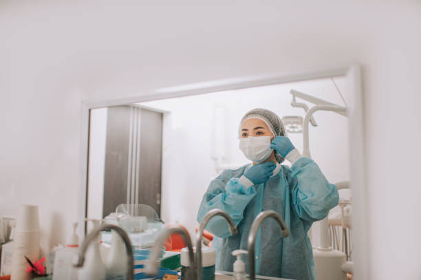 азиатские китайские женщины стоматолог носить защитную рабочую одежду сиз перед зеркалом в кабинете стоматолога готовится хирургии - hair net nurse scrubs asian ethnicity стоковые фото и изображения