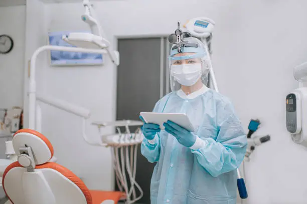 asian chinese dentist with PPE protective workwear holding digital tablet in dentist's office looking at camera