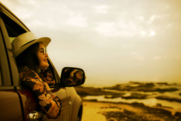 A close-up of a car reflecting a vibrant sunset on its windows, parked on a rural gravel road surrounded by fields and trees.