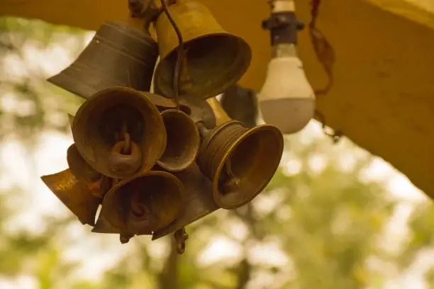 Photo of Group of tiny bronze bells hanging on a ceiling of a ancient hindu temple. Light emitting diode bulb in the blurred background.