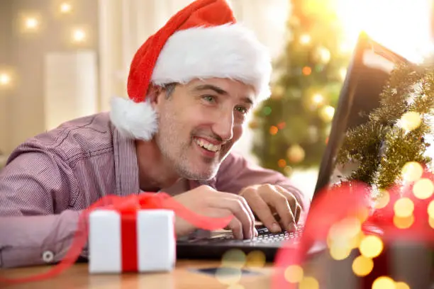 Photo of Smiling man with christmas hat using laptop at home