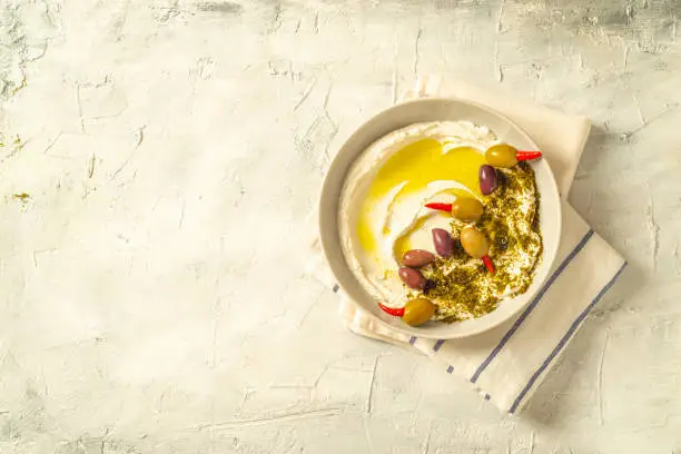 Photo of Popular middle eastern appetizer labneh or labaneh, soft white goat milk cheese with olive oil, hyssop or zaatar, olives on grey table, top view ,flat lay with copy space.