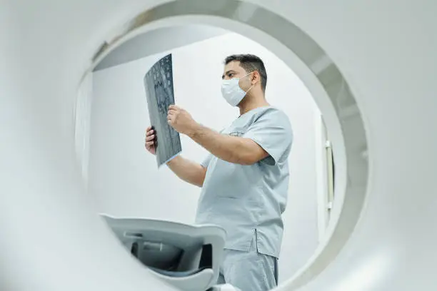 Serious mature mixed-race radiologist in mask and uniform looking at x-ray image of patient head in medical office environment in clinics