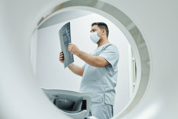 sério, radiologista maduro de máscara e uniforme olhando para a imagem de raio-x - radiologist - fotografias e filmes do acervo