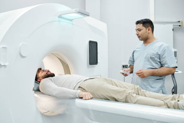 Young mixed-race male doctor in medical uniform touching button on control panel Young mixed-race male doctor in medical uniform touching button on control panel device in front of patient lying on long couchette mri scanner photos stock pictures, royalty-free photos & images