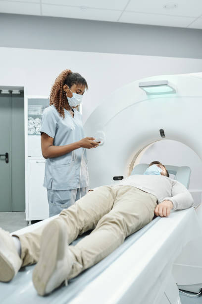 Young African female assistant in medical uniform and mask looking at patient Young African female assistant in medical uniform and mask touching button on control panel device in front of patient on long couchette cat scan stock pictures, royalty-free photos & images