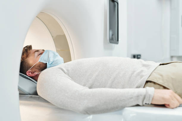 Young male patient in casualwear and protective mask lying on long couchette Young male patient in casualwear and protective mask lying on long couchette and moving into large medical equipment for examination pet scan photos stock pictures, royalty-free photos & images