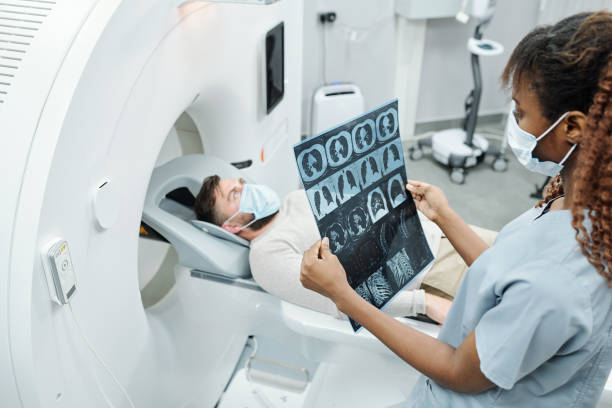 Young African radiologist in uniform and protective mask looking at x-ray image Young African radiologist in uniform and protective mask looking at x-ray image of patient lying on long couchette of medical equipment cat scan stock pictures, royalty-free photos & images